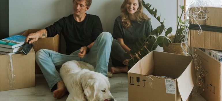 A man and a woman are happy that they settled in with Plano movers and are now sitting on the floor of their new home with their dog, surrounded by boxes.