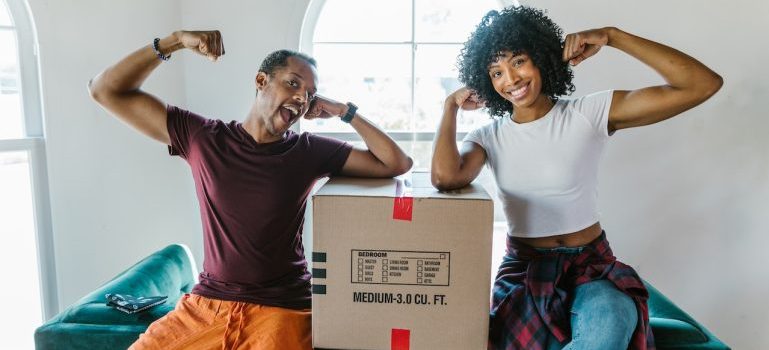 A man and a woman flexing their muscles after moving with Forth Worth movers and while sitting next to a box.